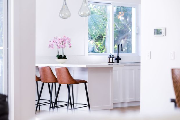 Bright, minimalist white kitchen with a peninsula breakfast bar, two rust-brown upholstered stools, white shaker-style cabinetry, pale benchtops, a black gooseneck tap, and a large window overlooking greenery. Two glass pendant lights and a small orchid add restrained decorative detail.
