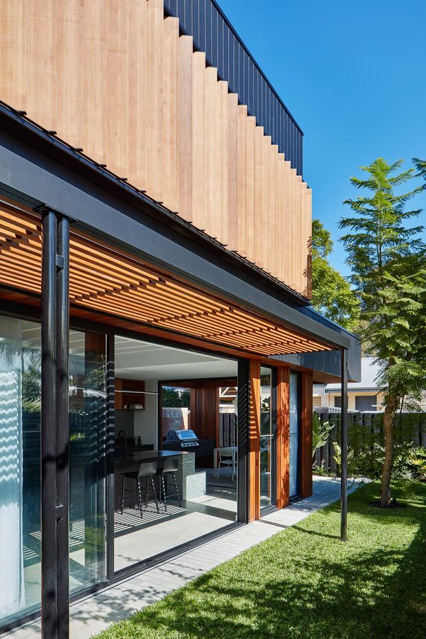 Exterior view of a contemporary multi-level residential extension with a dark metal-clad upper volume, stepped timber screening, extensive floor-to-ceiling glazing, and a covered outdoor entertaining area. The lower level opens to a compact kitchen or outdoor entertaining space with a stone island, bar stools, and built-in barbecue. Timber slatted soffits and warm timber-framed doors contrast with black structural framing. A landscaped lawn and established tree sit alongside the house.