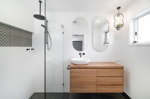 Bright contemporary bathroom with a frameless glass walk-in shower, black rain shower fittings, patterned decorative wall tile, floating timber vanity with white vessel basin, rounded mirrored cabinets, and a small pendant light. White walls and a dark floor create a clean, high-contrast finish.
