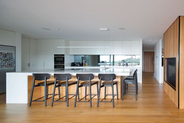 A bright, contemporary open-plan kitchen and dining area with a long white island and six dark bar stools. White full-height cabinetry, integrated appliances, a reflective splashback, timber flooring, and a warm timber-clad feature wall with a built-in fireplace define the space. Glazing beyond the kitchen provides a view outside.