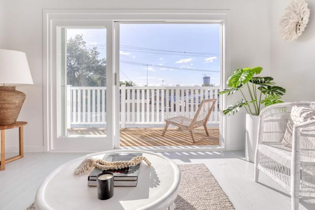 Bright, airy coastal-style living room with white walls and flooring, large glazed doors opening to a balcony or deck, a woven lounge chair, white wicker seating, a round light-toned coffee table, and indoor greenery. Strong natural sunlight creates a relaxed, inviting atmosphere.