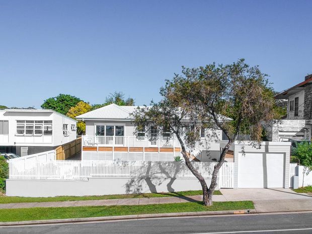 Street-facing residential exterior showing a white weatherboard-style house with a low pitched metal roof, broad windows, front balcony and white picket fencing. A separate garage and driveway sit to the right, with neighbouring homes and mature street trees visible.