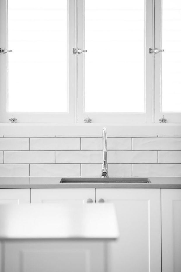 Bright monochrome kitchen detail featuring white shaker-style cabinetry, a grey countertop with an undermount sink and chrome gooseneck tap, white subway-tile splashback, and multiple white-framed windows above the bench. The foreground includes a blurred kitchen island or counter.