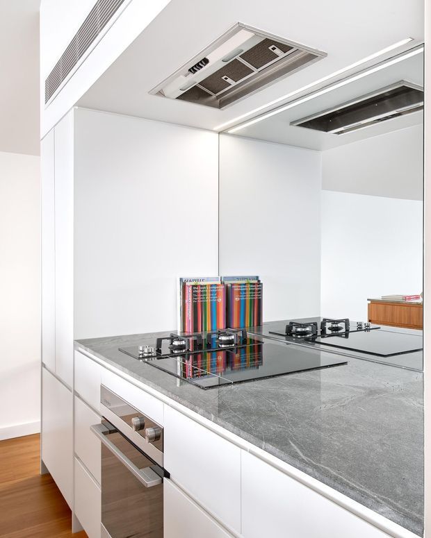 A bright, compact modern kitchen with white handleless cabinetry, a large grey stone island benchtop, black glass gas cooktop, stainless-steel integrated oven, mirrored splashback, and concealed ceiling ventilation. Timber flooring adds warmth, while linear recessed lighting and minimal detailing create a clean contemporary fit-out.
