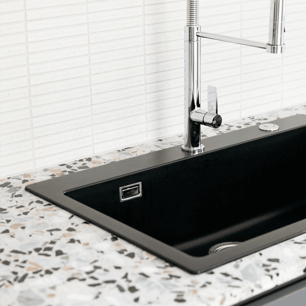 Close-up of a contemporary kitchen sink with a deep matte-black undermount basin, polished chrome pull-out faucet, and matching chrome controls. The sink is set into a terrazzo countertop with multicoloured stone aggregate, against a white horizontally tiled splashback.