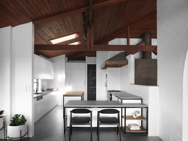 A bright, compact kitchen and dining area with white brick and painted walls, white cabinetry, dark grey flooring, and a prominent exposed timber structural ceiling. A small black-framed dining table with two chairs sits in the foreground, while a kitchen island, stainless-steel work surfaces, rangehood, and open shelving occupy the rear. Skylights bring natural light into the warm timber-lined ceiling.