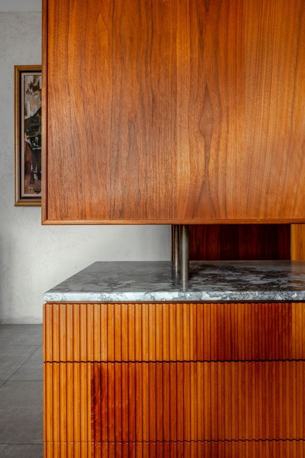 Close-up interior view of a richly grained warm timber kitchen or built-in joinery installation. The cabinetry features vertical fluted timber fronts and a large smooth timber overhead panel, paired with a dark grey veined stone benchtop and cylindrical metal supports. A pale textured wall and tiled floor are visible in the background.