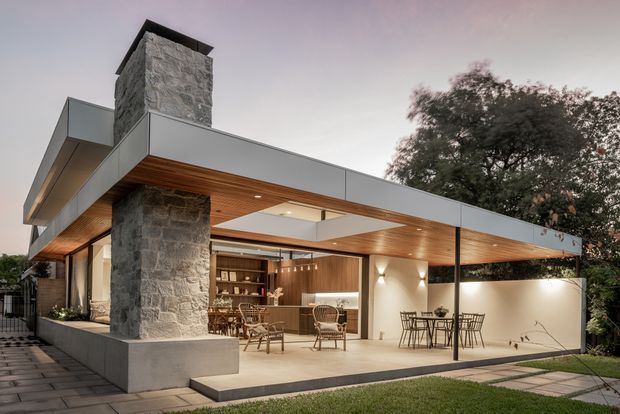Exterior architectural photograph of a contemporary single-storey home with a broad flat roof and deep covered entertaining terrace. The facade combines grey metal cladding, a prominent rough natural-stone chimney, warm timber soffit lining, expansive glazing, and sheltered outdoor dining and lounge areas. A built-in bar or kitchen is visible beneath the roof, with paved paths and landscaped planting around the house.