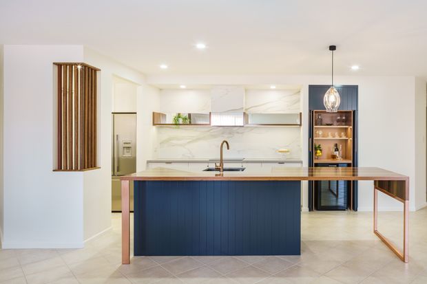 Bright contemporary kitchen with a large central island featuring dark navy-blue vertical-panel cabinetry, a pale stone benchtop, and warm timber or copper-toned supports. The rear kitchen has white marble-look splashback and cabinetry, integrated appliances, open timber shelving, and a dark blue built-in display or bar niche with glassware. A sculptural pendant hangs above the island, while a timber slatted feature screen is visible at left. The room is spacious, clean, and warmly lit.