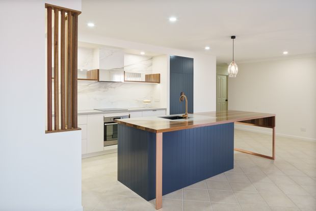 Bright open-plan kitchen and dining area featuring a large island with dark blue vertical-panel cabinetry, pale stone benchtops, copper-toned trim and legs, white cabinetry, and a marble-look splashback. A timber slatted divider, pendant light, recessed ceiling lighting, and tiled flooring complete the refined contemporary fit-out.