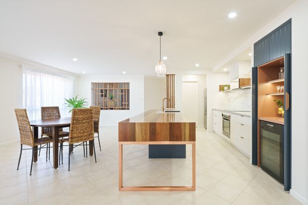 Bright open-plan kitchen and dining area with white cabinetry, pale tiled flooring, a timber island bench, woven dining chairs, and a dark-framed built-in display and beverage cabinet. A timber slatted divider and glazed shelving add visual texture to the otherwise clean, contemporary interior.