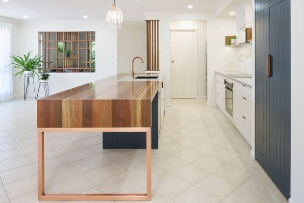 Bright contemporary kitchen with a large timber-topped island and waterfall-style end detail, white cabinetry, pale tiled flooring, a dark blue vertical-panel pantry or storage wall, and timber slatted screening around a window. A pendant light, integrated appliances, and indoor plants complete the clean, open-plan space.