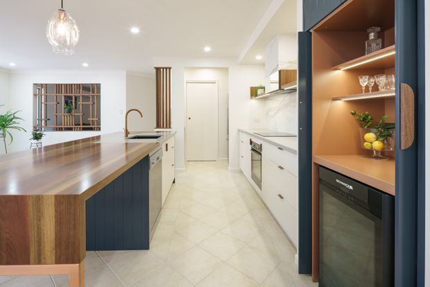Bright contemporary kitchen featuring a large timber-topped island with dark blue vertical-panel cabinetry, white perimeter cabinetry, pale tiled flooring, integrated appliances, and a tall illuminated display cabinet with warm timber shelving and glassware. A decorative timber screen and indoor plants add natural detail.