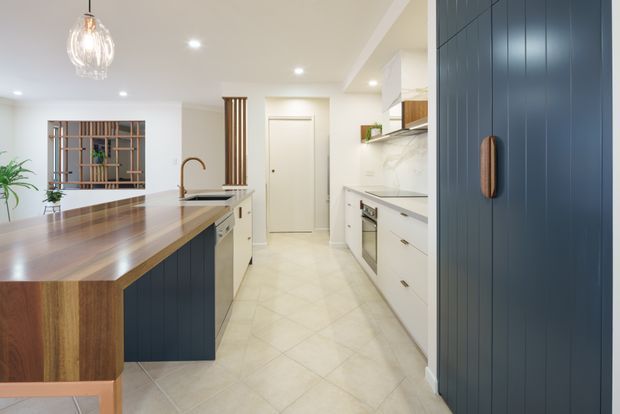 Bright contemporary kitchen featuring a large timber island with dark navy-blue panelled cabinetry, white perimeter cabinetry, pale tiled flooring, integrated stainless-steel appliances, a brass-toned tap, and a decorative pendant light. A vertical timber screen and glazed shelving detail add warmth and texture.