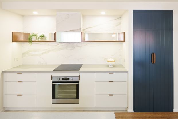 A compact contemporary kitchen with white flat-panel cabinetry, a pale stone-look benchtop and matching marble-look splashback, integrated cooktop and stainless-steel oven. Open timber shelves with plants and small decor sit beneath a concealed rangehood, while a tall navy-blue paneled cabinet with timber handles provides a strong contrasting feature. Light flooring and recessed ceiling lights create a bright, clean space.