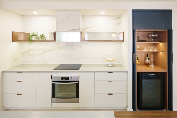 A bright, contemporary galley-style kitchen with white marble-look wall surfaces, white handleless cabinetry with slim timber pulls, an integrated cooktop and oven, and open timber display shelving. A dark blue built-in drinks cabinet with illuminated timber shelving and a wine refrigerator forms a strong feature at the right. The space has recessed ceiling lighting and a warm timber floor area.