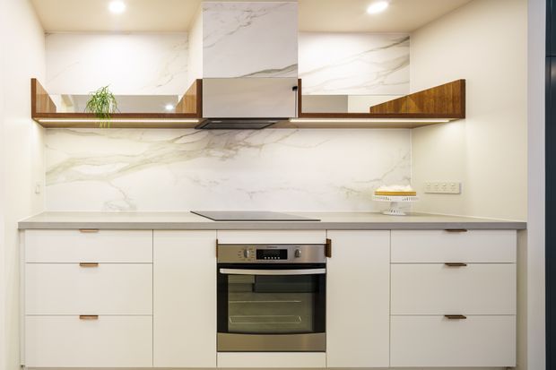 Bright contemporary kitchen with white cabinetry, a pale marble-look splashback and matching rangehood surround, integrated oven and cooktop, and warm timber open shelving and accents. Recessed ceiling lights and a clean linear layout create a refined, compact cooking space.