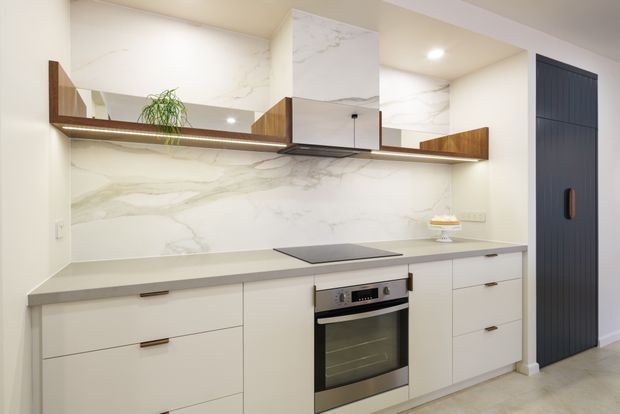 Contemporary compact kitchen with pale handleless-style cabinetry, a grey stone benchtop, a large marble-look splashback and integrated electric oven. A timber open shelf with mirror backing runs along the wall, while a dark vertically panelled door provides contrast. Recessed ceiling lighting and minimal detailing create a clean, refined fit-out.