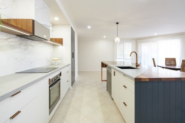Bright contemporary kitchen with white cabinetry, a large central island, pale stone surfaces, marble-look splashback, timber accents, and a dark blue island base. The open-plan space extends toward a dining area with sheer curtains and abundant natural light.