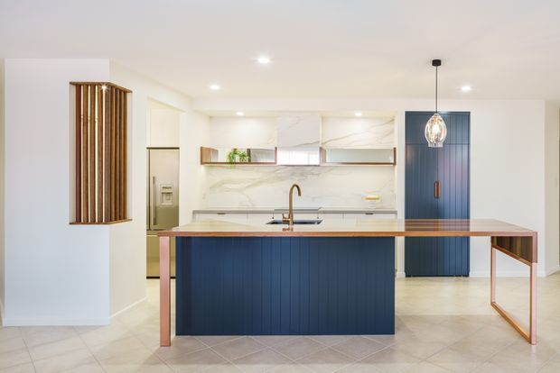 Bright contemporary kitchen with a large central island featuring deep navy-blue vertical-panel cabinetry, a pale stone benchtop, and warm timber edging. The rear wall has a white marble-look splashback, open timber-framed shelving, white cabinetry, and integrated appliances. A matching navy pantry or utility door, warm timber accents, pendant lighting, and pale tiled flooring complete the clean, refined scheme.