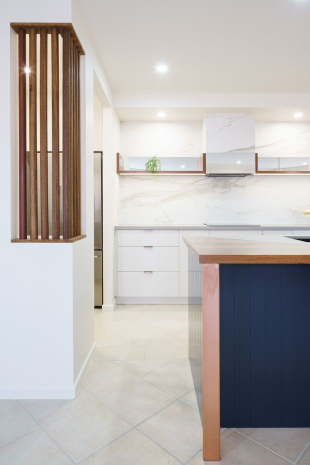 Bright contemporary kitchen viewed from the adjoining circulation area, featuring white cabinetry, a large island with a dark navy panelled base and timber trim, marble-look splashback and benchtops, integrated rangehood, and pale tiled flooring. A vertical timber slat screen forms a decorative architectural divider on the left.