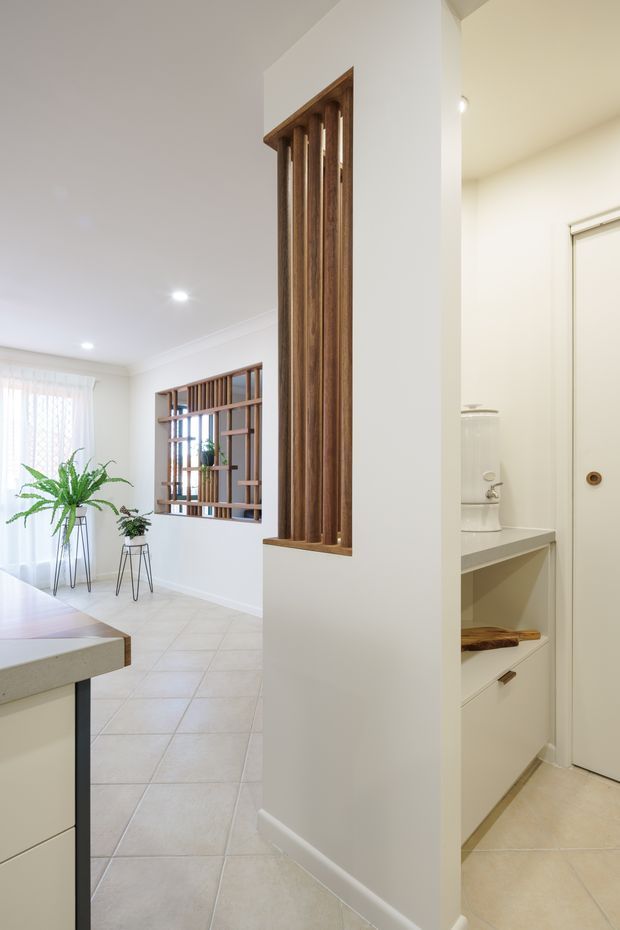 Bright residential interior showing a tiled open-plan transition between a kitchen and living/dining area. White walls and cabinetry are complemented by warm timber vertical screens and timber-framed internal windows. A partially visible bathroom or utility area with a toilet and built-in shelving opens on the right.