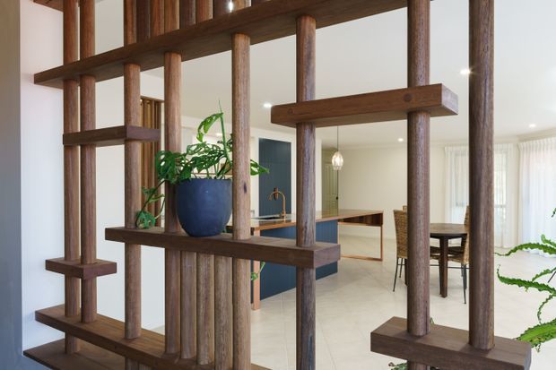 Interior view of a contemporary open-plan home, framed by a substantial dark timber vertical screen with integrated shelves and potted greenery. Beyond it is a light-toned kitchen with a dark island, timber cabinetry, pendant lighting, and an adjacent dining area with woven dining chairs. Pale tiled flooring and white walls create a bright, warm setting.