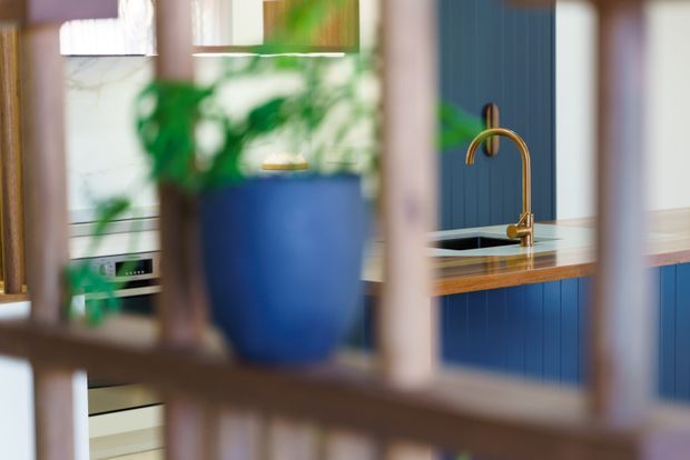 A contemporary kitchen viewed through a blurred timber screen or balustrade. The kitchen features deep blue vertically panelled cabinetry, a warm timber benchtop, a black sink, and a curved brass/gold tap. White appliances and pale walls provide contrast, while a large blue planter with greenery is softly out of focus in the foreground.