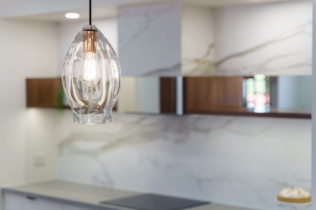 Close-up view of a refined contemporary kitchen, focused on a sculptural clear-glass pendant light with a copper-toned fitting. Behind it are white marble-look slab surfaces, dark timber-toned cabinetry or feature panels, mirrored splashback elements, and a dark stone-look benchtop with an integrated cooktop.