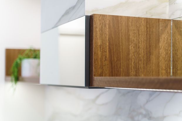 Close-up view of a contemporary bathroom vanity or built-in cabinet, featuring warm wood-grain cabinetry, dark trim, and white marble-look wall surfaces. A mirrored cabinet section reflects a bright, minimalist interior with a small plant.