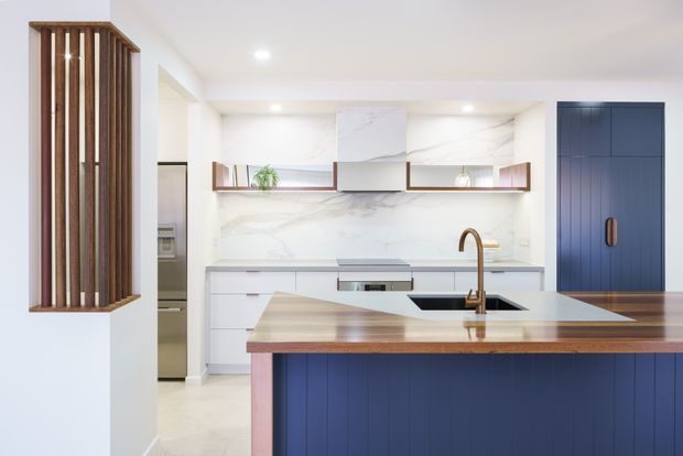 Bright contemporary kitchen featuring a large island with a timber benchtop and dark blue vertically panelled cabinetry, white cabinetry, marble-look wall surfaces, open timber-framed shelving, brass tapware, and a tall blue pantry or storage cabinet. A vertical timber screen adds architectural detail at the left.