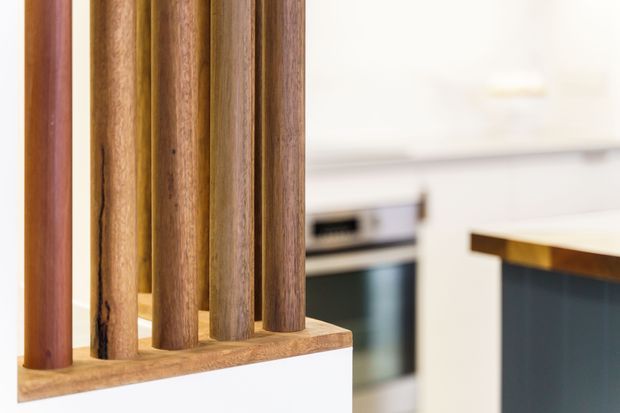 Close-up interior view of a contemporary kitchen featuring a vertical timber slat divider or balustrade in the foreground. A white island with a dark blue-green base and timber-edged benchtop is visible to the right, with white cabinetry and integrated stainless-steel appliances in the background.