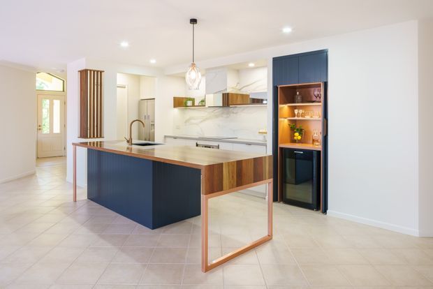 Bright contemporary kitchen with a large island featuring deep blue panelled cabinetry, warm timber edging and an open timber-framed seating area. The kitchen includes a pale stone benchtop, marble-look splashback, integrated appliances, a dark blue built-in bar or beverage cabinet with open shelving and a wine fridge, plus light tiled flooring and white walls. An entry hall and decorative vertical timber screen are visible in the background.