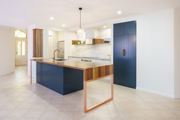 Bright contemporary kitchen with a large island featuring deep navy blue cabinetry, warm timber accents, and a pale stone benchtop. The room has light tiled flooring, white walls and ceiling, marble-look splashback panels, integrated appliances, a tall navy pantry or storage door, recessed downlights, and a pendant light. A hallway and entry door are visible beyond the kitchen.