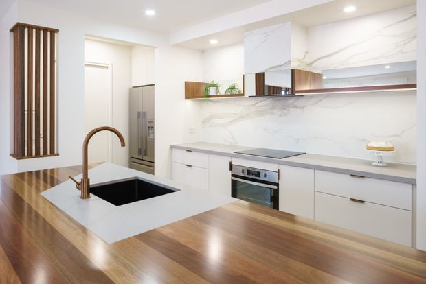 A bright contemporary kitchen with a large timber island, white cabinetry, integrated stainless-steel appliances, a deep black sink, and a curved bronze-toned tap. The island and cabinetry are topped with pale grey engineered-stone surfaces, while a dramatic white marble-look splashback extends across the cooking wall. Vertical timber slats and open timber shelving add warmth and texture.