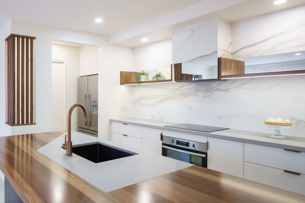 Bright contemporary kitchen featuring a large island with a dark undermount sink and brushed brass gooseneck tap, white waterfall-style benchtop, integrated dark cooktop, stainless-steel oven, extensive white cabinetry, and a dramatic white veined stone splashback. Warm timber flooring and timber accents add contrast, while a vertical timber slat screen and mirrored upper detail provide architectural interest.