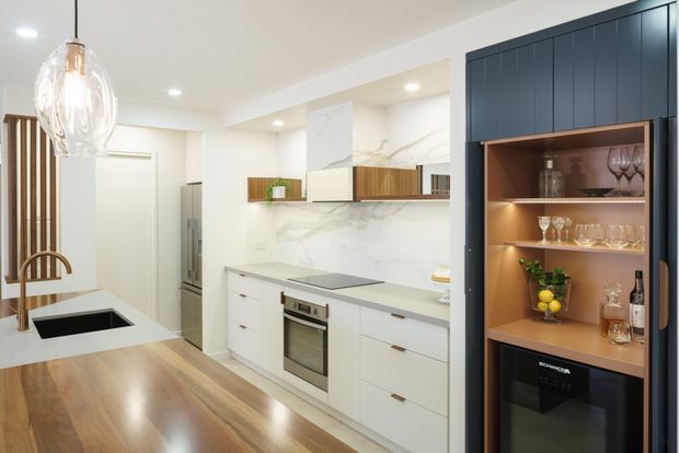 Bright contemporary kitchen featuring white flat-panel cabinetry, a pale stone-look splashback and benchtops, integrated stainless-steel appliances, warm timber flooring, and a dark navy built-in bar or display cabinet with illuminated timber shelving. A dining area and sculptural pendant light are visible at the left.