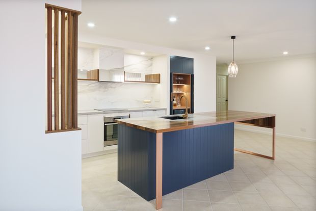 Bright contemporary open-plan kitchen and dining area with a large island featuring deep blue vertical-panel cabinetry, warm timber edging, and a pale stone benchtop. White cabinetry and a marble-look splashback line the rear wall, with integrated appliances and an illuminated timber display niche. A timber vertical screen partially separates the kitchen from the adjacent area. Light tiled flooring, recessed ceiling lights, and a pendant add a clean, refined finish.