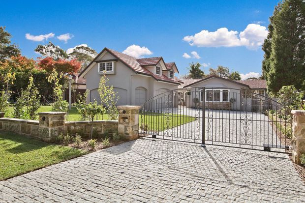 A sunny suburban residential property viewed from the street, featuring a rendered multi-gabled house with tiled pitched roofs, a covered entry, detached or attached garage, decorative metal driveway gates, stone masonry pillars and retaining walls, and a cobblestone driveway. Mature landscaped gardens surround the home.
