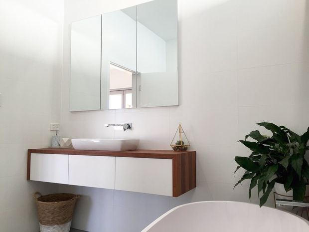 Bright contemporary bathroom featuring a wall-mounted timber vanity with white flat-panel cabinetry, a white vessel basin, chrome wall-mounted tapware, and a large mirrored cabinet. White tiled walls, a freestanding bath edge, and a leafy plant create a clean, airy feel.