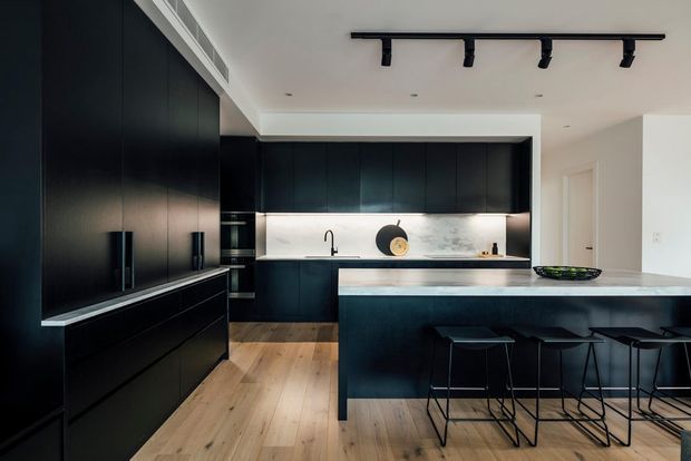 Contemporary kitchen with extensive matte dark cabinetry, a large island with integrated seating, a pale stone benchtop and backsplash, timber flooring, and recessed track lighting. The streamlined black-and-white palette is balanced by warm wood flooring.