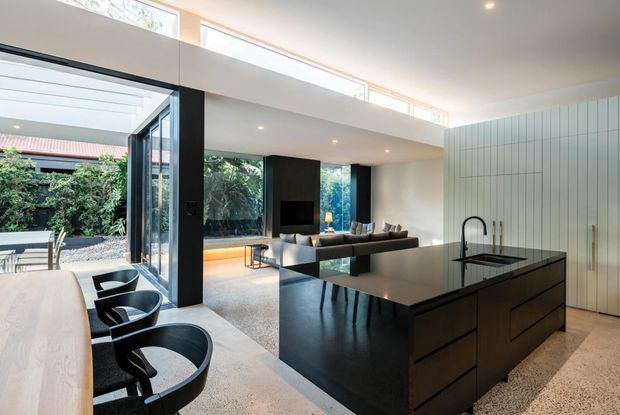 A bright, open-plan contemporary kitchen and living area with a large black island, integrated sink and dark cabinetry, adjacent to a dining table and black chairs. The space features polished concrete or terrazzo-like flooring, extensive glazing, a double-height void, clerestory windows, and a partially visible courtyard garden beyond the glass.