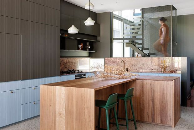 Contemporary open-plan kitchen with a large timber island and integrated seating, dark vertically grooved cabinetry, pale blue-grey lower cabinetry, and a pink-toned stone splashback. Pendant lights hang above the island, while a stairway and glazed interior partition are visible in the background.