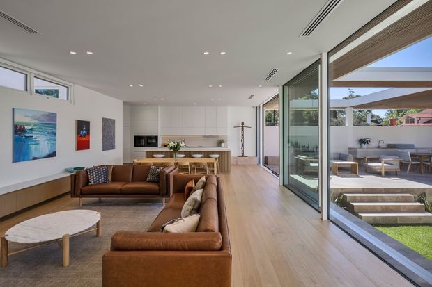 Bright open-plan contemporary interior combining a living room, dining area and minimalist kitchen, with full-height sliding glass doors opening to a landscaped outdoor courtyard and dining terrace. The room features warm timber flooring, white built-in kitchen cabinetry, leather sofas, recessed lighting and extensive indoor-outdoor connection.