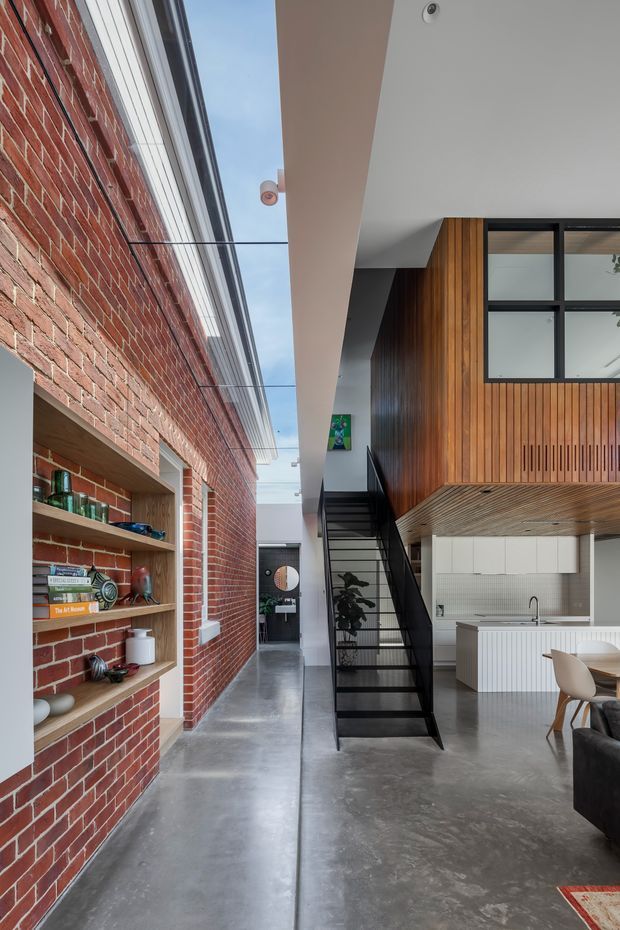 A tall, contemporary open-plan interior showing a polished concrete floor, exposed red-brick wall with recessed shelving, a black steel stair, and a compact white kitchen beneath a mezzanine clad in vertical timber. Large black-framed glazing, a high ceiling, and a long skylit circulation zone create an airy industrial-residential atmosphere.