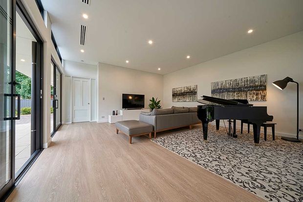 Bright, spacious contemporary living room with pale timber flooring, full-height black-framed glazing opening to an outdoor area, a grey sectional sofa, ottoman, black grand piano, floor lamp, television, and abstract wall art. Recessed ceiling lighting and linear air vents contribute to a clean, refined interior.