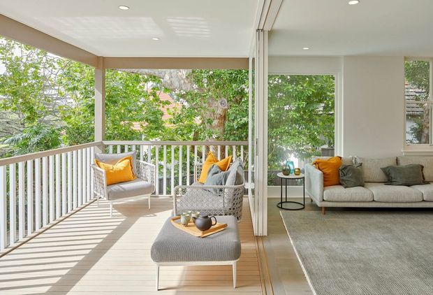 Bright, airy indoor-outdoor living space with a covered timber-lined balcony or terrace opening directly into a contemporary living room through wide sliding or folding glazed doors. The terrace has white railings, wicker lounge chairs, and an ottoman, while the living room contains a light neutral sofa, grey rug, and warm mustard cushions. Abundant greenery is visible outside, and recessed ceiling lights provide a clean modern finish.