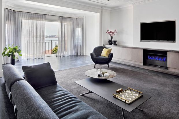 A spacious, refined living room with a large grey sectional sofa, black coffee tables, a round lounge chair, and a wall-mounted television above a built-in linear fireplace and cabinetry. White decorative wall mouldings and sheer full-height curtains frame glazed doors opening to a balcony, while a dark grey rug and polished grey floor create a sophisticated monochromatic palette.