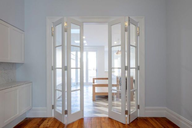 Bright, refined interior view from a kitchen or adjacent room through a set of white-framed glazed bi-fold French doors into a sunlit dining area. The foreground has polished warm timber flooring, pale blue-grey walls, white cabinetry, decorative skirting and ceiling mouldings, while the adjoining room contains a timber dining table and patterned floor tiles.