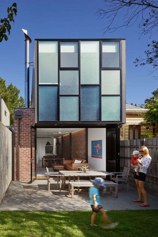 Exterior view of a narrow, multi-storey urban home extension with a projecting upper level clad in a grid of translucent textured glazing and dark metal framing. The lower level opens onto a paved outdoor dining and lounge area, with exposed red brick walls, timber fencing, and a tall metal flue. Adjacent traditional residential buildings and greenery surround the contemporary addition.